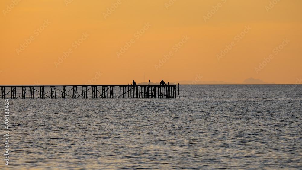 Silhouetted people sitting and relaxing on a long wharf jetty over ocean water against a beautiful golden orange sunset sky in Raja Ampat, West Papua, Indonesia
