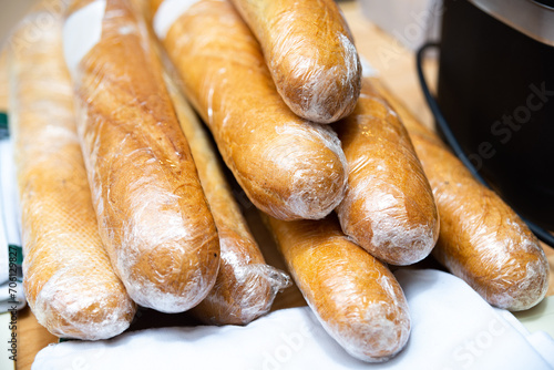 Fresh Baguettes Wrapped in Plastic on Table. Multiple freshly baked baguettes wrapped in cling film, ready for sale or storage on a wooden tabletop.