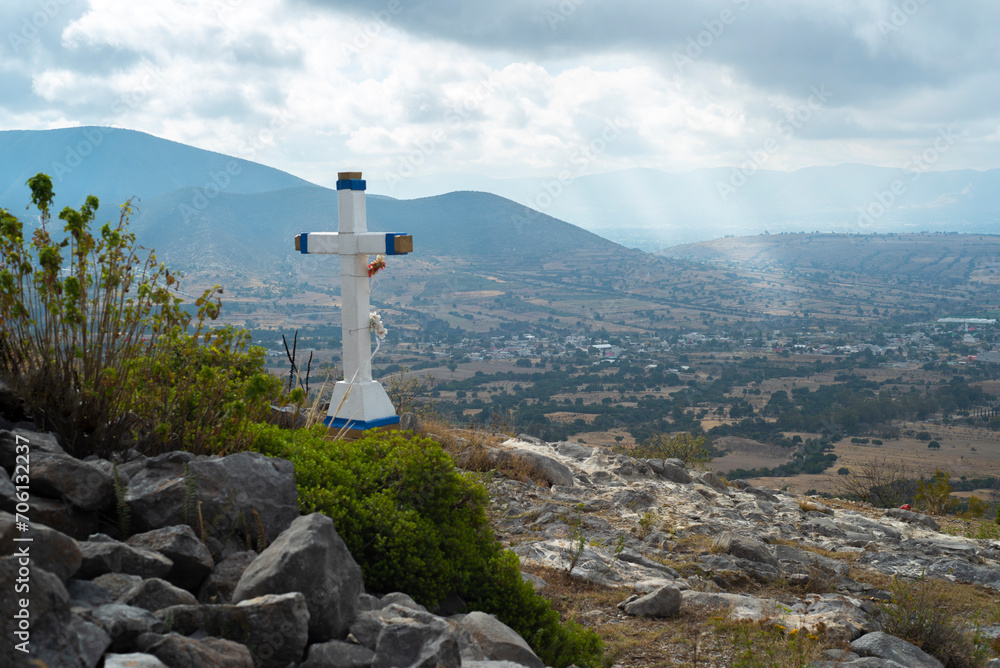 Paisaje. Cruz blanca cristiana sobre cerro. mirando al pueblo con montañas y cielo nublado con rayos de luz de fondo.