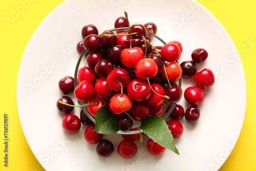 Plate and glass bowl with sweet cherries on yellow background