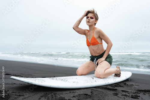 Woman surfer posing on her knees on white surfboard  looking at camera on black sand beach on background of ocean waves during summer holiday. Authenticity female surfer dressed in bikini top  shorts