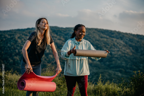Black and Caucasian women chatting happily while carrying yoga mats for an outdoor session