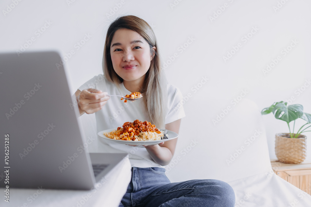 Asian woman eating pasta while watching series from notebook, sitting on white bed and working at home, smiling and looking at camera. Asian woman eating pasta while watching series from notebook, sitting on white bed and working at home, smiling and looking at camera.