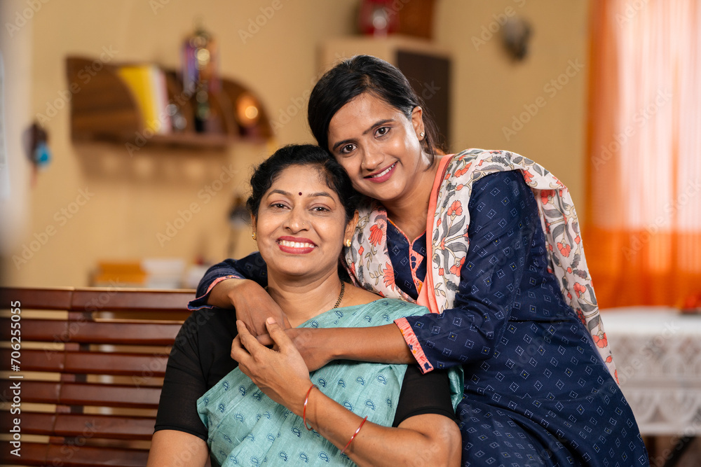happy smiling Mother with daughter looking at camera by embracing or hugging each other at home - concept of family support, companion and emotional bonding.