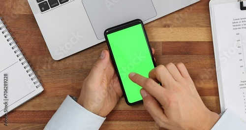 Man moves index finger across green blank screen of modern smartphone at workplace top view photo