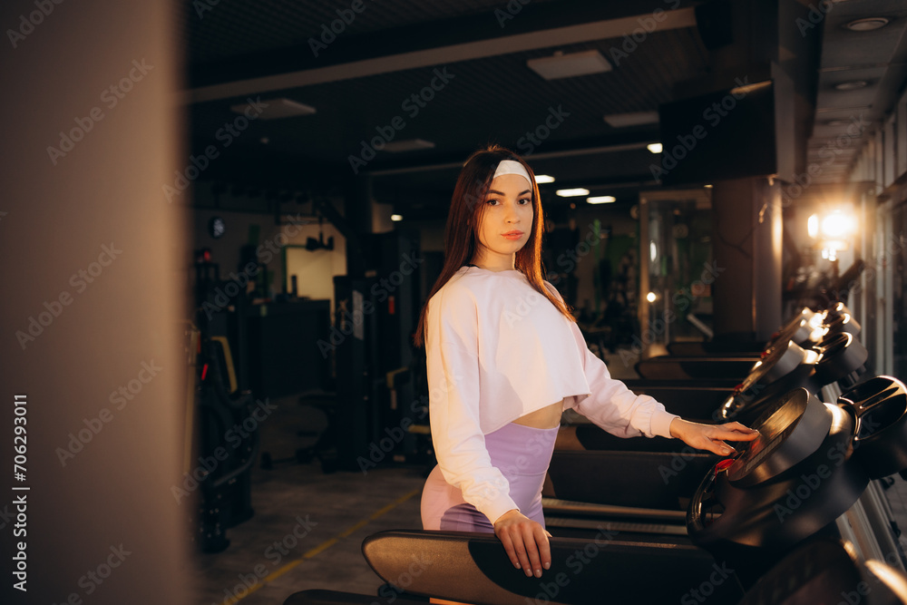 young sports girl in pink sportswear stands in the gym near the treadmills, an attractive woman trains in a dark fitness room young sports girl in pink sportswear stands in the gym near the treadmills, an attractive woman trains in a dark fitness room