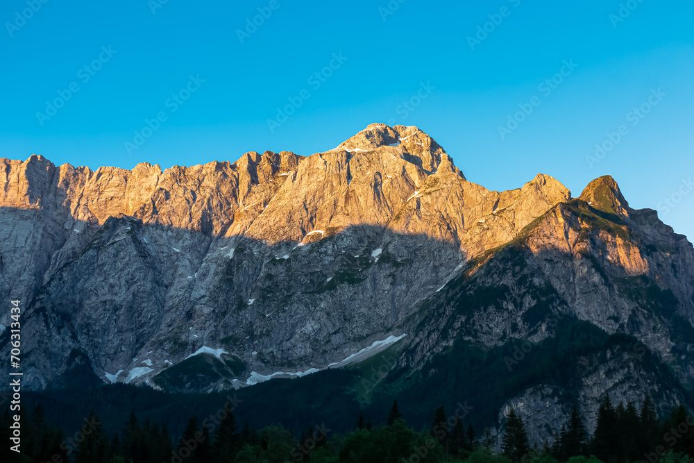 Panoramic sunrise view of mountain summit Mount Mangart in Julian Alps, Tarvisio, Friuli Venezia Giulia, Italy, Europe. Rock walls in warm red orange colors. Majestic landscape at alpine Lake Fusine