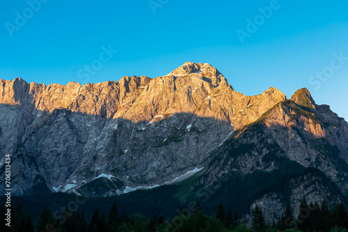 Panoramic sunrise view of mountain summit Mount Mangart in Julian Alps, Tarvisio, Friuli Venezia Giulia, Italy, Europe. Rock walls in warm red orange colors. Majestic landscape at alpine Lake Fusine