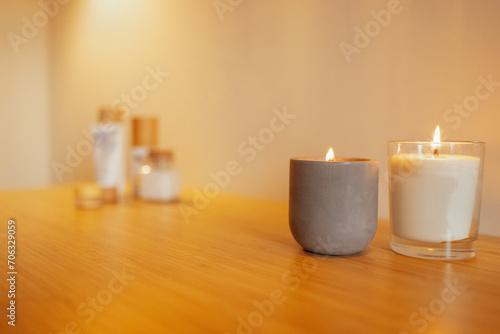 Two lighted candles in a ceramic and glass candlestick on a wooden table. Cosmetic and beauty products in the background.