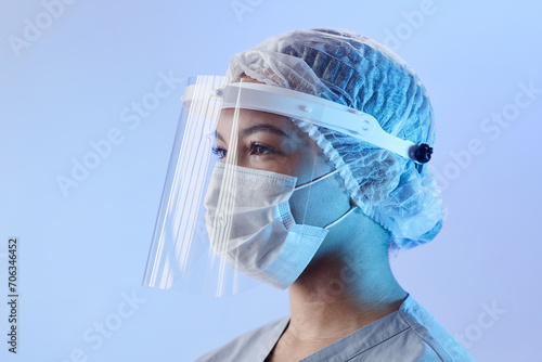 Medium close up with side view of serious woman doctor in medical face shield, mask and sterile hat looking away standing at pastel blue background photo