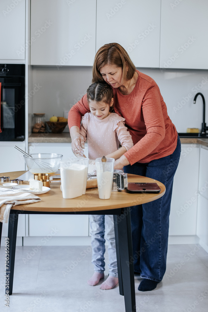 A 60-year-old Caucasian woman kneads dough with her hands with an 8-year-old girl in a bright kitchen, there are various kitchen utensils on the table A 60-year-old Caucasian woman kneads dough with her hands with an 8-year-old girl in a bright kitchen, there are various kitchen utensils on the table