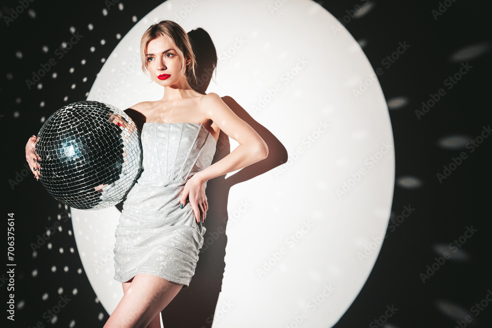 Naklejka premium Young beautiful smiling blond female in trendy evening silver dress. Sexy carefree woman posing near white wall in studio in a circle of light. Fashionable model with bright makeup. Holds disco ball