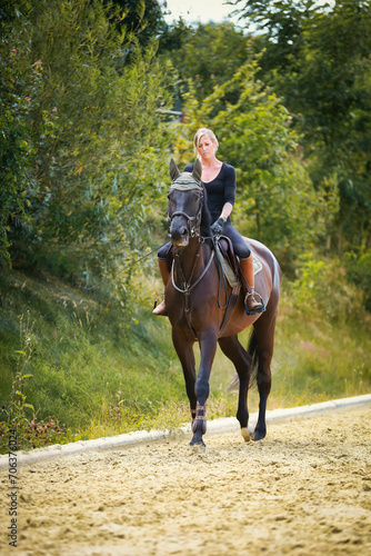 Young blonde girl woman with horse on the riding arena in summer.