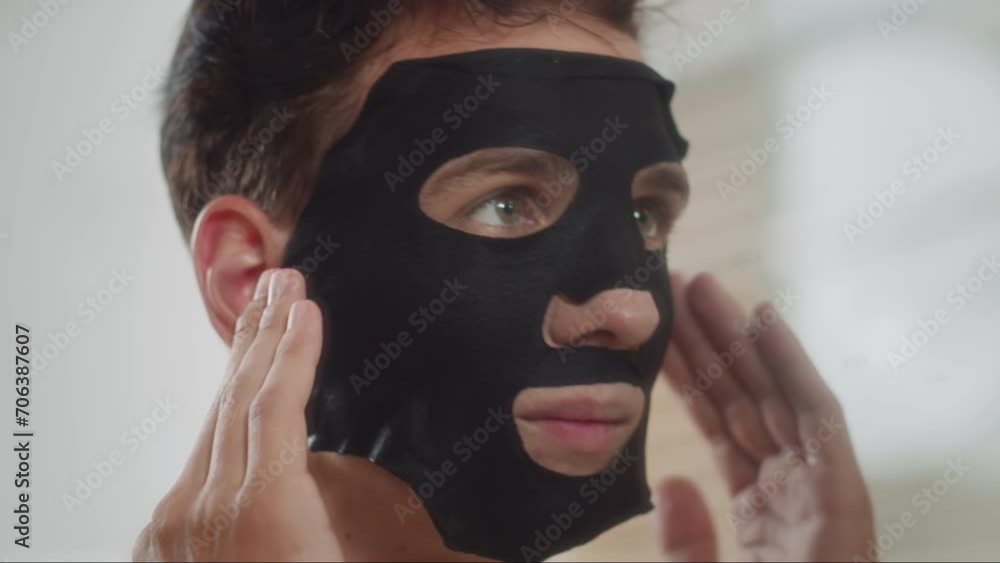 Big close up of young man of mixed ethnicity using black hydrating mask on face during skin care procedures in front of mirror in bathroom Big close up of young man of mixed ethnicity using black hydrating mask on face during skin care procedures in front of mirror in bathroom