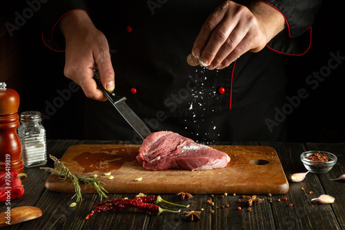 Chef sprinkles salt on raw fresh meat. Preparing lamb steak before baking. Working environment in a restaurant or hotel kitchen.