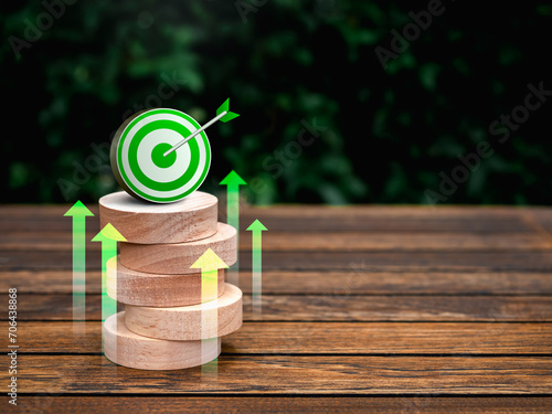 3d green target icon on the top of round wooden blocks stack on green natural leaves background with copy space. Green business target development, goal and success, growth marketing plan concepts.