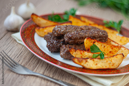 Lula kebabs on a white plate with a red border with baked potatoes and greens.