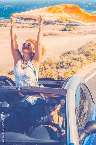 Happy women traveler enjoy road trip with a convertible car. One woman outside the roof have fun. Scenic desert destination. Car driving. Excited couple of friends in a vehicle on travel. Adventure © simona