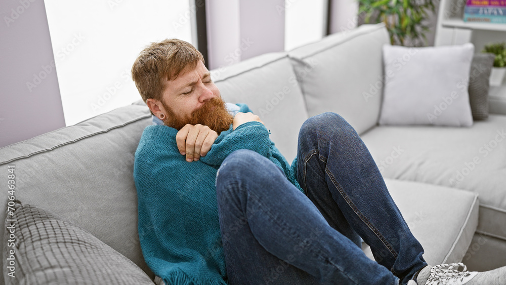 Chilly winter's tale, handsome young redhead man freezing, finds cozy comfort in a warm blanket, sitting on sofa, battling the cold indoors at home.