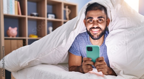 Young hispanic man using smartphone lying on bed at bedroom