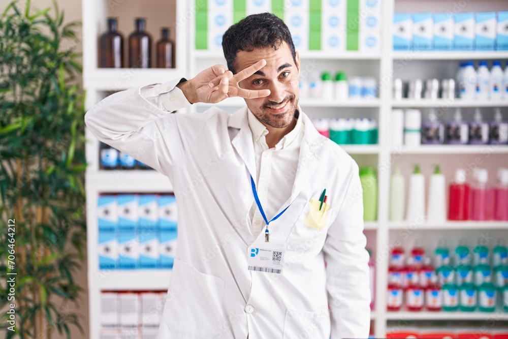 Handsome hispanic man working at pharmacy drugstore doing peace symbol with fingers over face, smiling cheerful showing victory Handsome hispanic man working at pharmacy drugstore doing peace symbol with fingers over face, smiling cheerful showing victory