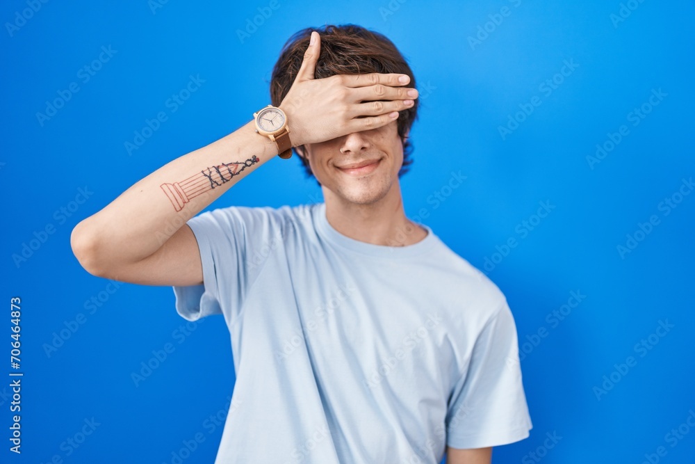 Hispanic young man standing over blue background smiling and laughing with hand on face covering eyes for surprise. blind concept. Hispanic young man standing over blue background smiling and laughing with hand on face covering eyes for surprise. blind concept.