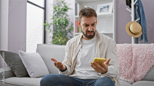 Young, handsome hispanic man with beard sitting upset on sofa at home, using smartphone. serious adult male texting, engrossed in technology indoors.