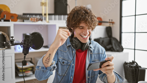 A jubilant young man with headphones in a music studio celebrates good news on his smartphone.