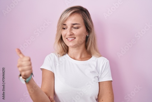 Young blonde woman standing over pink background looking proud, smiling doing thumbs up gesture to the side © Krakenimages.com