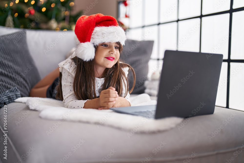 Fototapeta premium Adorable hispanic girl having video call lying on sofa by christmas tree at home