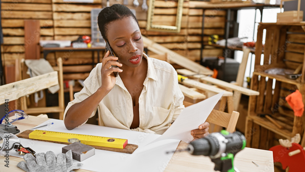 Attractive african american woman carpenter in serious conversation on smartphone, reading important document amidst her carpentry workshop Attractive african american woman carpenter in serious conversation on smartphone, reading important document amidst her carpentry workshop
