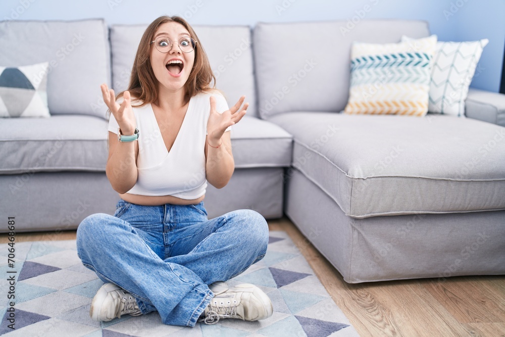 Young caucasian woman sitting on the floor at the living room celebrating crazy and amazed for success with arms raised and open eyes screaming excited. winner concept Young caucasian woman sitting on the floor at the living room celebrating crazy and amazed for success with arms raised and open eyes screaming excited. winner concept