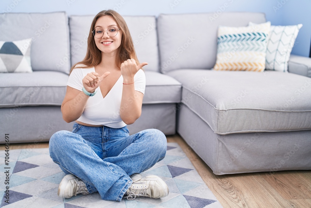 Young caucasian woman sitting on the floor at the living room pointing to the back behind with hand and thumbs up, smiling confident Young caucasian woman sitting on the floor at the living room pointing to the back behind with hand and thumbs up, smiling confident