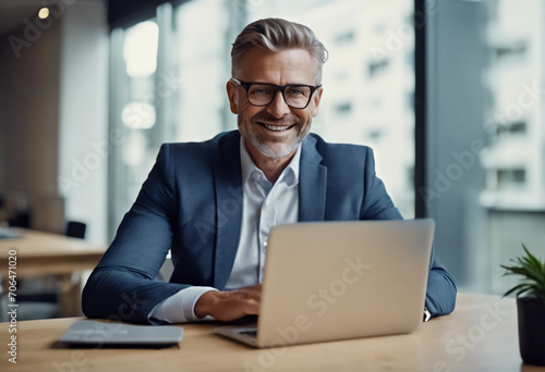 Eleganza Professionale- Affascinante Business Man di Mezza Età, Sorridente, Seduto al Desk con il Laptop in un Ufficio Moderno photo