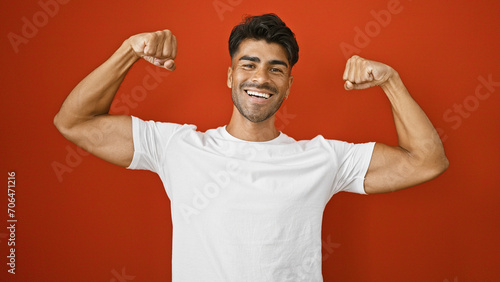 A cheerful young hispanic man flexes his muscles confidently against an isolated red background, exuding strength and positivity.
