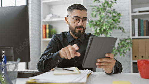 A bearded young man in glasses uses a tablet while working in a modern office, portraying focus and professionalism.