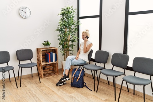 Young blonde woman talking on smartphone sitting on chair at waiting room