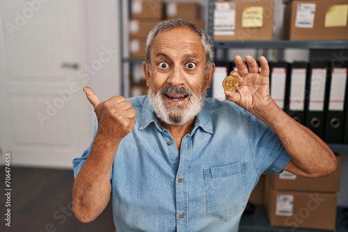 Cheerful senior man showing thumbs up, holding a bitcoin coin in office, smiling wide with open mouth, pointing to the side with joy and confident optimism.