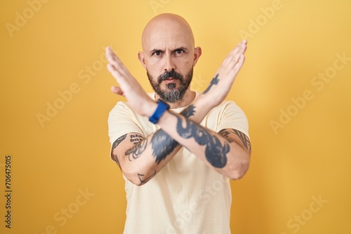 Hispanic man with tattoos standing over yellow background rejection expression crossing arms doing negative sign, angry face