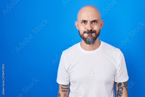 Hispanic man with tattoos standing over blue background puffing cheeks with funny face. mouth inflated with air, crazy expression. photo