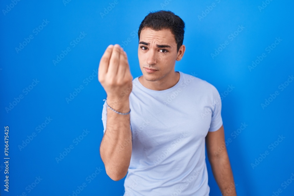 Young hispanic man standing over blue background doing italian gesture with hand and fingers confident expression Young hispanic man standing over blue background doing italian gesture with hand and fingers confident expression
