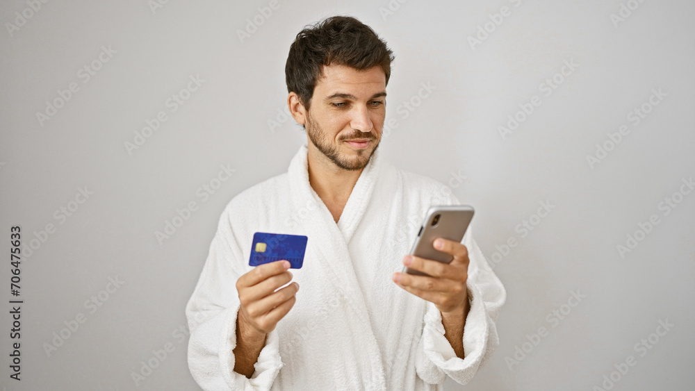 Handsome hispanic man in white bathrobe holding credit card and smartphone against isolated background. Handsome hispanic man in white bathrobe holding credit card and smartphone against isolated background.