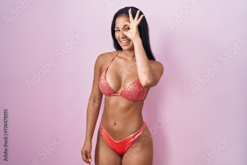 Hispanic woman wearing bikini doing ok gesture with hand smiling, eye looking through fingers with happy face.