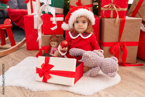 Adorable blonde toddler unpacking gift sitting by christmas tree at home