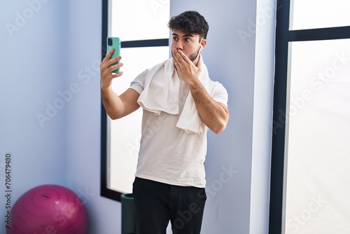 Hispanic man with beard doing video call with smartphone at the gym covering mouth with hand, shocked and afraid for mistake. surprised expression