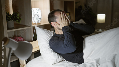 Bald hispanic man with beard, stressed in bedroom at night, covers face with his hands, lamps on.