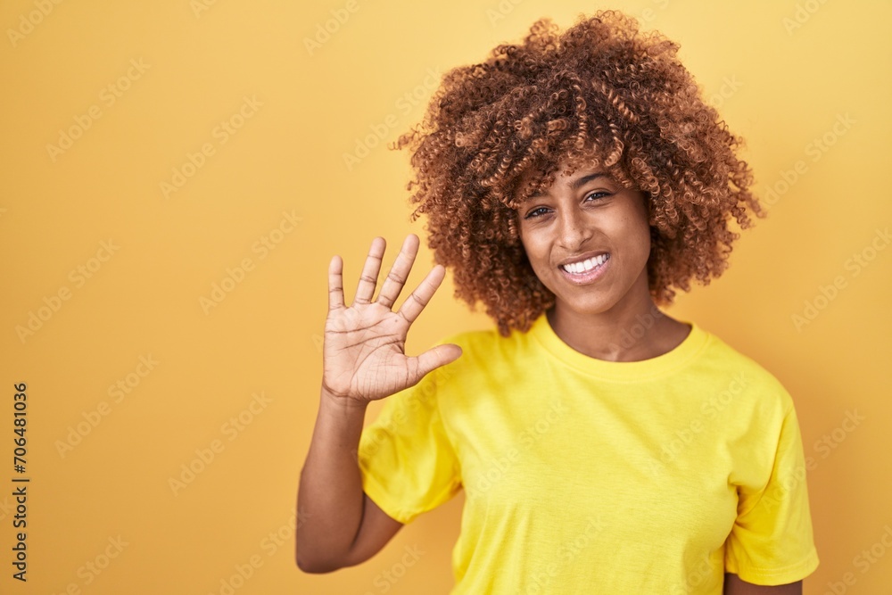 Young hispanic woman with curly hair standing over yellow background showing and pointing up with fingers number five while smiling confident and happy. Young hispanic woman with curly hair standing over yellow background showing and pointing up with fingers number five while smiling confident and happy.