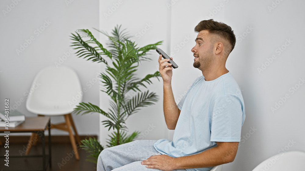Young man with beard in casual attire using smartphone indoors in a minimalist room with plants.