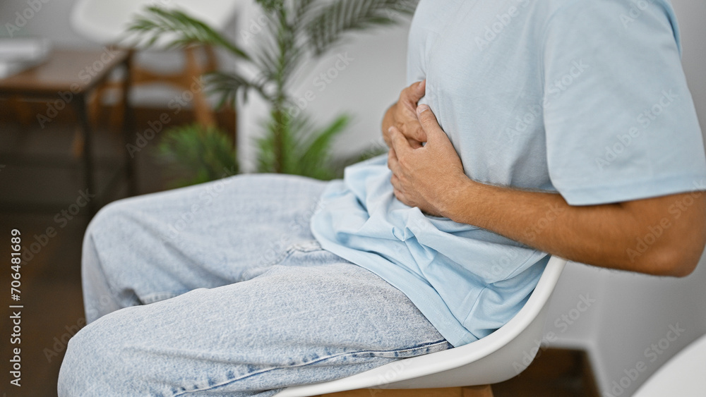 Hispanic man with stomach pain sitting in a modern indoor waiting room. Hispanic man with stomach pain sitting in a modern indoor waiting room.