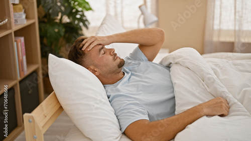 A young man with a beard showing discomfort lying in bed within a cozy bedroom setting. © Krakenimages.com